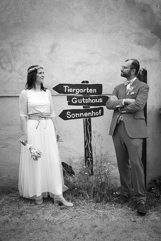Schwarz-weiß-Bild eines Brautpaares vor einem gemalten Wegweiser auf Gut Boltenhof